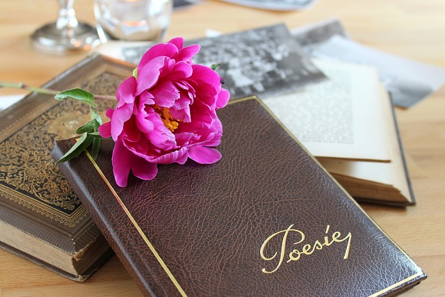 A leather-bound poetry book with the word "Poesie" on the cover, adorned with a vibrant pink peony flower resting on top, alongside other old books in the background